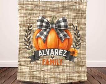 May include: A fall-themed blanket featuring an orange pumpkin with a black and white checkered bow. The design includes a banner with the text "ALVAREZ FAMILY" and a sunflower. The background has a burlap texture.