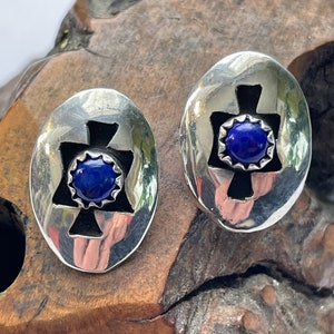 May include: A pair of silver earrings with a blue gemstone in the center of each earring. The earrings are oval shaped and have a cross design.