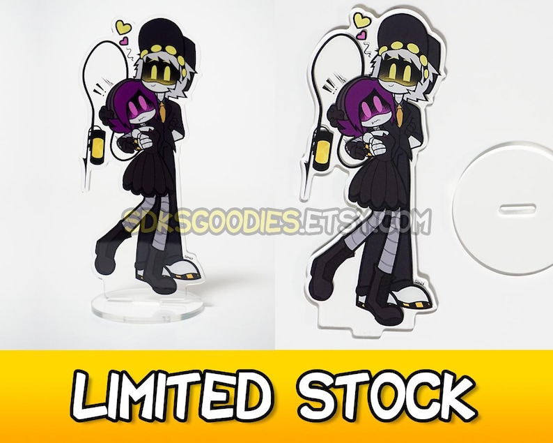 May include: Acrylic stand featuring a cartoon couple in black outfits, with yellow accents and hearts. The stand has a clear base and a white background. The text "LIMITED STOCK" is at the bottom.