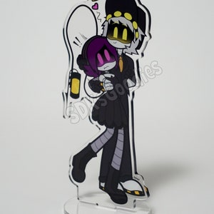 May include: A clear acrylic stand-up featuring a cartoon couple embracing. The characters are in black outfits with yellow and purple accents. One character wears a hat, and the other has purple hair. A lightbulb and hearts are drawn above them.