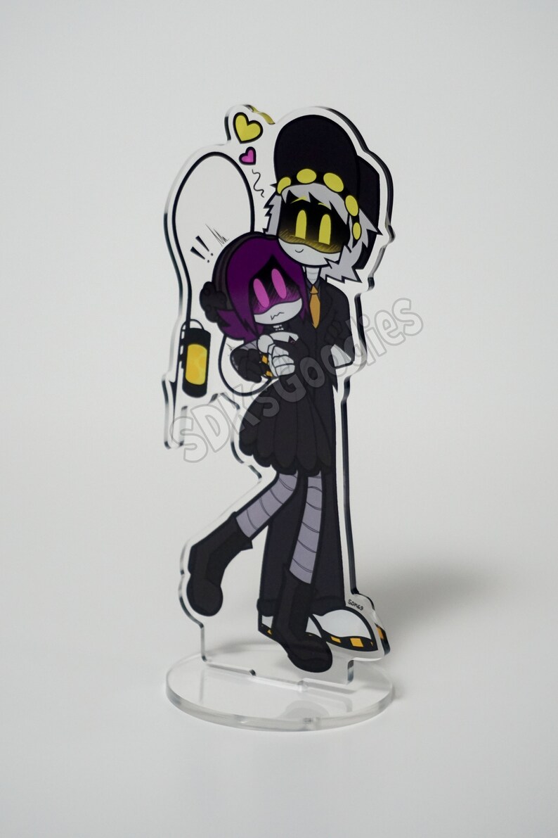 May include: A clear acrylic stand-up featuring a cartoon couple embracing. The characters are in black outfits with yellow and purple accents. One character wears a hat, and the other has purple hair. A lightbulb and hearts are drawn above them.