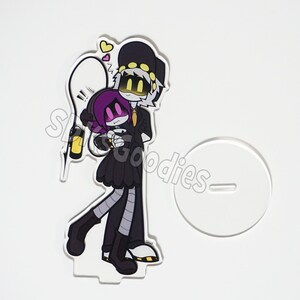 May include: A clear acrylic stand featuring a cartoon illustration of two characters, one with a black hat and the other with a purple head. The characters are hugging. A white stand is to the right. The text "SLS Goodies" is visible.