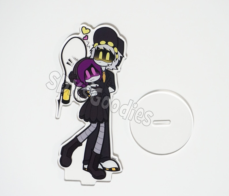 May include: A clear acrylic stand featuring a cartoon illustration of two characters, one with a black hat and the other with a purple head. The characters are hugging. A white stand is to the right. The text "SLS Goodies" is visible.