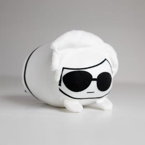 Peut inclure: Un jouet en peluche blanc en forme de personnage de dessin animé, avec des lunettes de soleil noires et une coiffure blanche. Le jouet a un corps arrondi avec des lignes noires et de petits pieds blancs.