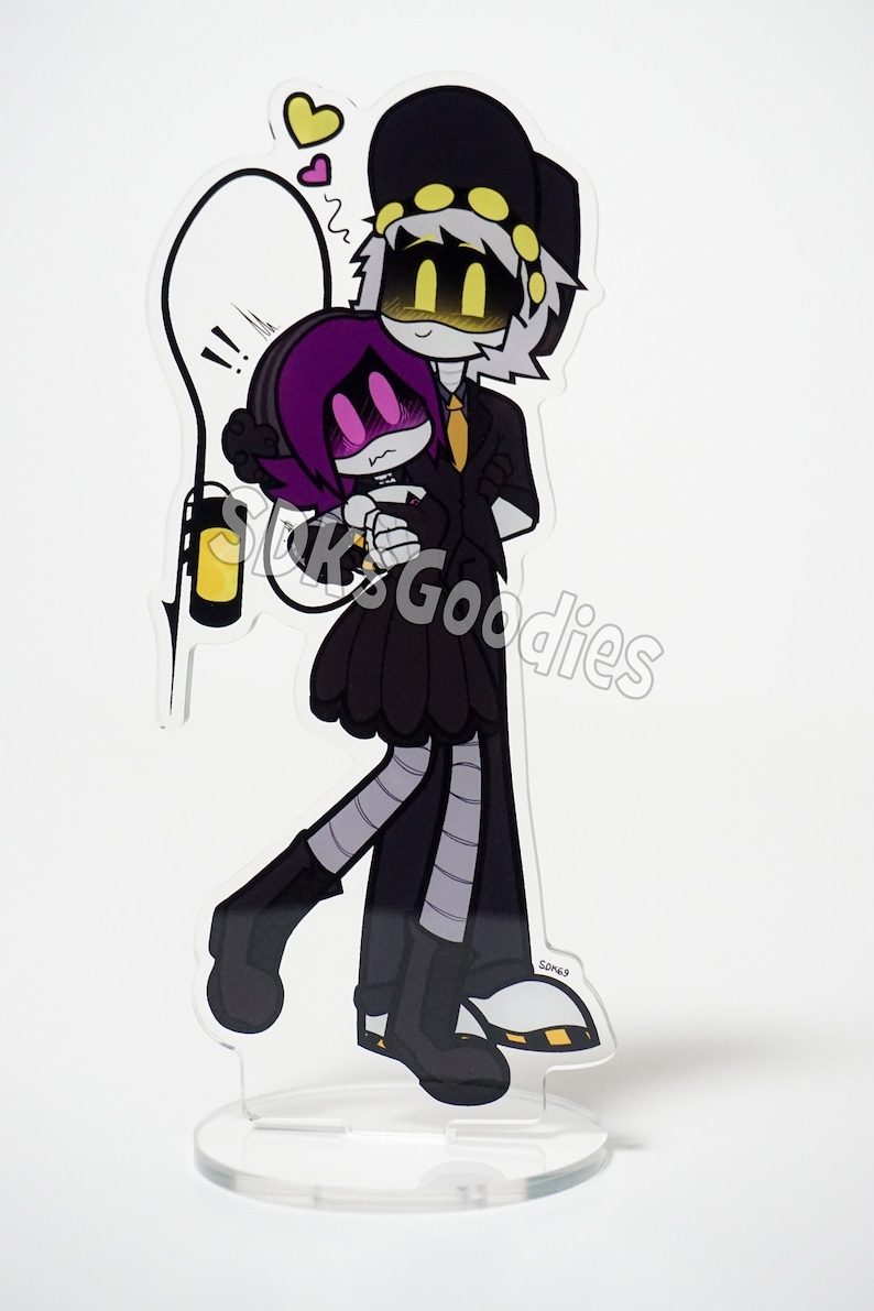 May include: A clear acrylic stand-up featuring a cartoon couple embracing. The characters are in black, white, and purple, with yellow accents. One character wears a black hat and suit, while the other has purple hair and a black dress. A yellow lantern is also included.