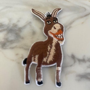 May include: Embroidered patch featuring a cartoon donkey with a brown body, tan and white accents, and a wide grin. The patch has a white border and is designed for application on clothing or accessories.