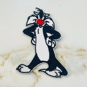 May include: An embroidered iron-on patch featuring a cartoon black cat with a red nose. The cat is standing with its arms crossed and has a mischievous expression.