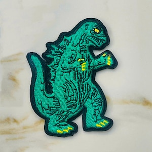 May include: Green embroidered iron-on patch of a Godzilla-like monster with yellow eyes and claws.