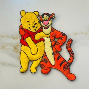 May include: Embroidered patch featuring Winnie the Pooh and Tigger. Pooh is wearing a red shirt and Tigger is wearing his signature orange and black stripes.