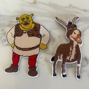 May include: Two embroidered patches featuring Shrek and Donkey from the Shrek movie franchise. Shrek is green and wearing a brown vest and red pants. Donkey is brown and has a white stripe down his back.