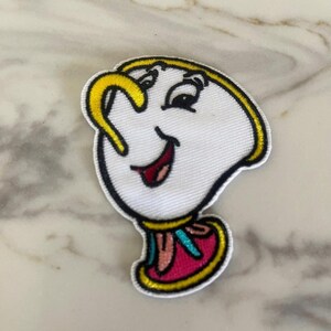 May include: A white embroidered patch of the character Lumiere from Disney's Beauty and the Beast. Lumiere is a teapot with a yellow handle and a smiling face. He is wearing a pink and blue sash.