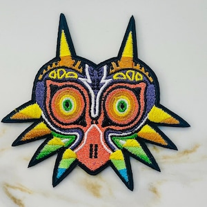 May include: A colorful embroidered patch featuring the Majora's Mask from the Legend of Zelda video game series. The mask is a stylized, demonic face with large, yellow eyes, a red mouth, and a purple and green design.