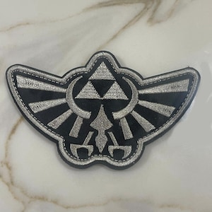 May include: A black and silver embroidered patch featuring the Triforce symbol from the Legend of Zelda video game series. The Triforce is surrounded by a winged design.