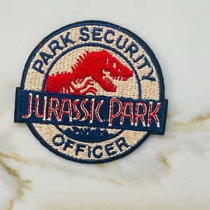May include: A blue and white embroidered patch with the words "Park Security Jurassic Park Officer" and a red dinosaur silhouette.