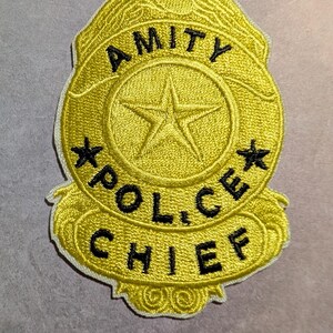 May include: Embroidered yellow patch with the words "AMITY POLICE CHIEF" in black lettering. The patch has a badge shape with a star in the center and two smaller stars on either side. The patch is outlined in white.