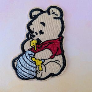 May include: Embroidered patch featuring a cartoon bear in a red shirt, eating honey from a blue and white striped pot. The bear is beige with black outlines. Yellow honey drips from the bear's mouth and the pot.