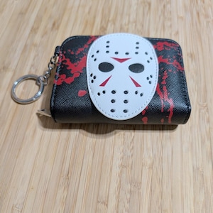 May include: Black wallet with a white hockey mask design and red splatter accents. The wallet has a silver key ring attached. The mask has black eye holes and red details. The wallet is on a wooden surface.