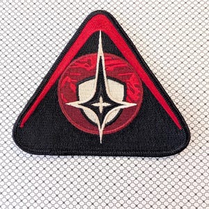 May include: A black, triangular patch with a red border and a central design. The design features a red circle with a white and black emblem, and a white starburst. The patch is likely an emblem or insignia.