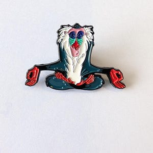 May include: Enamel pin featuring a cartoon baboon in a seated yoga pose. The pin has a dark teal body, red hands and feet, and a white beard. The baboon's face has blue, pink, and red details. The pin is a collectible item.
