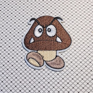 May include: Embroidered patch featuring a brown mushroom-like character with an angry expression. The character has white eyes, a beige stem, and two brown feet. The patch is outlined in white and set against a patterned background.