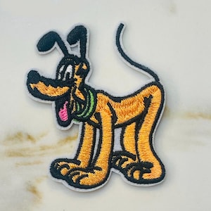 May include: An embroidered iron-on patch featuring a cartoon dog with black ears, a green collar, and a pink tongue. The dog is brown and white and is standing on all fours.