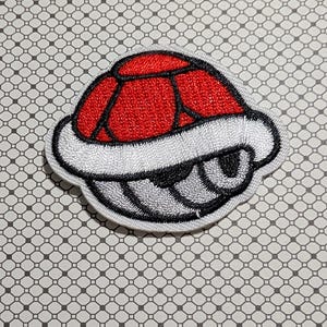 May include: Embroidered patch featuring a red and black turtle shell design with a white border. The shell has a segmented pattern and a gray underside. The patch is on a patterned background.