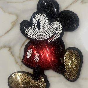 May include: A red, white, and gold sequined iron-on patch of Mickey Mouse. The patch is approximately 4 inches tall and 3 inches wide.