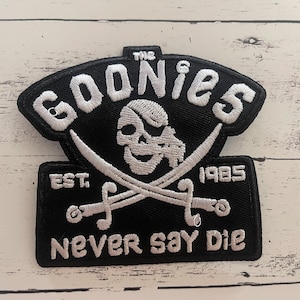 May include: Black and white embroidered patch featuring the text "THE GOONIES" above a skull and crossed swords. Below, the patch reads "EST. 1985" and "NEVER SAY DIE". The patch is a rectangular shape with a curved top.