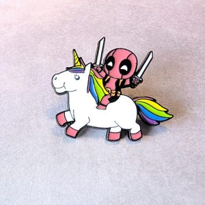 May include: A colorful enamel pin featuring a white unicorn with a rainbow mane and tail. A pink character with two swords rides the unicorn. The pin has a pink, yellow, and blue color scheme.