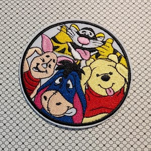 May include: Embroidered circular patch featuring Winnie the Pooh, Tigger, Piglet, and Eeyore. The characters are in a group hug, with vibrant colors and detailed stitching. The patch has a white border and is set against a patterned background.