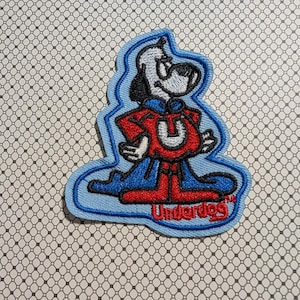 May include: Embroidered patch featuring the cartoon character Underdog. The patch has a light blue border and depicts a white dog with black ears and a red and blue superhero outfit. The word "Underdog" is in red text.