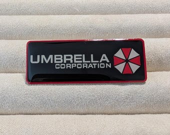 May include: A rectangular black and red badge with the text "UMBRELLA CORPORATION" in white. The badge features a red and white umbrella logo on the right side. The badge is glossy and appears to be made of metal or plastic.