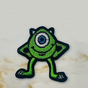 May include: Green one-eyed monster with black outlines and white teeth, standing with its arms raised and legs spread apart. The monster has a blue eye with a white pupil.