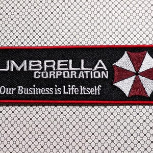 May include: A rectangular black patch with the text "UMBRELLA CORPORATION" in white, and "Our Business is Life Itself" below. A red and white umbrella logo is on the right. The patch has a red border.