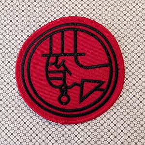 May include: A round, red patch with a black border and a black symbol in the center. The symbol is an abstract design with curved and angular lines. The patch is on a patterned background.