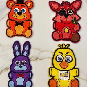 May include: Four embroidered patches featuring characters from the popular video game Five Nights at Freddy's. The patches are in the shape of a bear, a fox, a rabbit, and a chicken. The bear is orange and brown, the fox is red, the rabbit is purple, and the chicken is yellow. The patches are all about 3 inches tall.