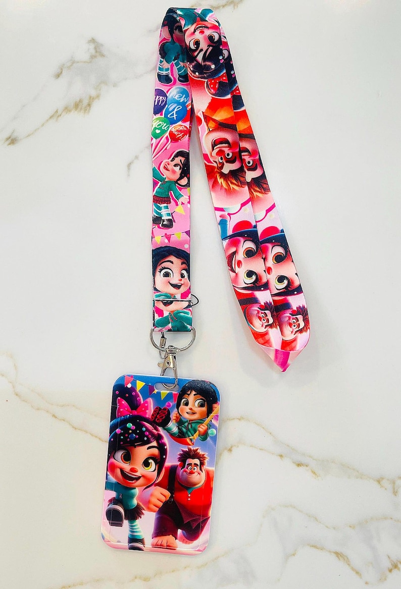 May include: A pink and blue lanyard with a cartoon image of characters from the movie Wreck-It Ralph. The lanyard has a matching ID card holder with the same characters. The text on the lanyard reads "Happy New & You".