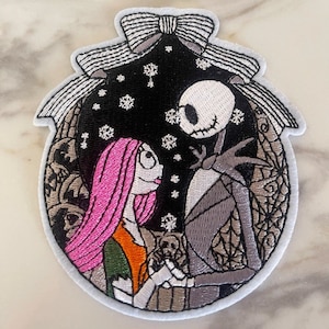 May include: Embroidered patch featuring Jack Skellington and Sally from The Nightmare Before Christmas. The patch is white with a black and white image of the characters in a snowy scene. The characters are holding hands and looking at each other.