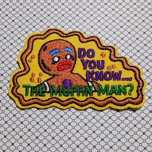 May include: Embroidered patch featuring a gingerbread man with blue eyebrows and red lips. The patch has the text "Do You Know... The Muffin Man?" in green and purple on a yellow background with a brown border.