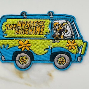May include: A blue and green embroidered patch of the Mystery Machine van from Scooby-Doo. The van has the words "The Mystery Machine" written on the side. There are three figures inside the van, two with blonde hair and one with brown hair.