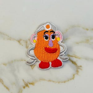 May include: Embroidered iron-on patch featuring a cartoon character with orange skin, white eyes, pink lips, and a white hat with a yellow circle. The character has pink ears and a purple nose.