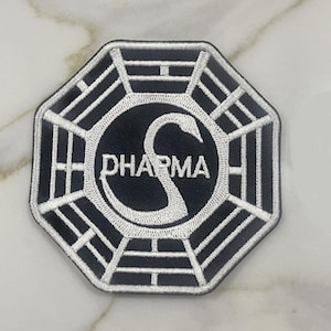 May include: Black and white embroidered patch with an octagonal shape. The patch features a circular design with the word "DHARMA" and a stylized "S" in the center. The outer border is white, and the background is black.