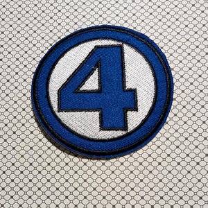 May include: A circular blue and white embroidered patch featuring a large blue number "4" inside a white circle, surrounded by a blue border. The patch has a black stitched edge and is likely an emblem or logo.