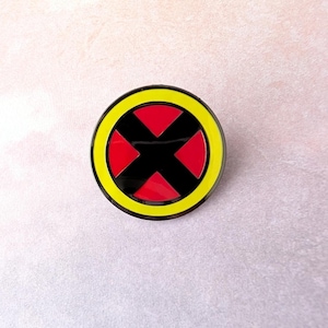 May include: A round pin featuring the X-Men logo. The pin has a black X in the center, surrounded by red triangles, a yellow ring, and a black border. The pin is set against a light pink background.