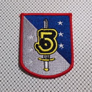 May include: Embroidered patch with a shield shape, featuring a blue and silver background. A yellow number 5 is centered, with a sword design and three white stars. The patch has a red border.