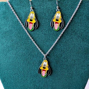 May include: A silver-toned necklace and matching earrings set. Each piece features a yellow, black, and green enamel pendant in the shape of a cartoon dog's head with a pink tongue. The earrings hang from silver hooks.