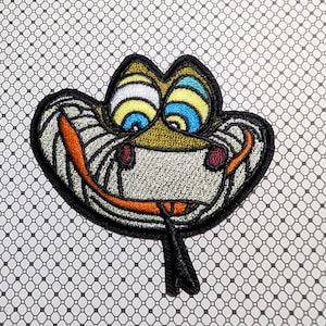 May include: Embroidered patch featuring a cartoon snake with a wide grin and a long, black tongue. The snake has yellow, blue, and white eyes, and orange and red accents. The patch is outlined in black and has a gray and white body.