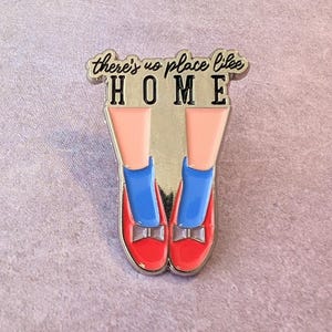 May include: Enamel pin featuring red shoes with bows, blue socks, and pink legs. The pin has the words "there's no place like HOME" in a cursive font above the shoes. The pin has a silver border.
