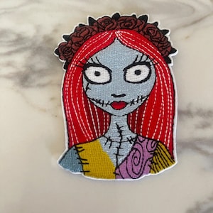 May include: Embroidered iron-on patch featuring a cartoon character with red hair, a stitched-together dress, and a crown of roses. The character has a stitched-together face with large eyes and a stitched-together mouth.