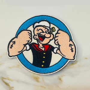 May include: An embroidered iron-on patch featuring a cartoon image of Popeye the Sailor Man with his signature red and black sailor outfit, white hat, and a pipe in his mouth. He is flexing his biceps and smiling. The patch is surrounded by a blue circle.